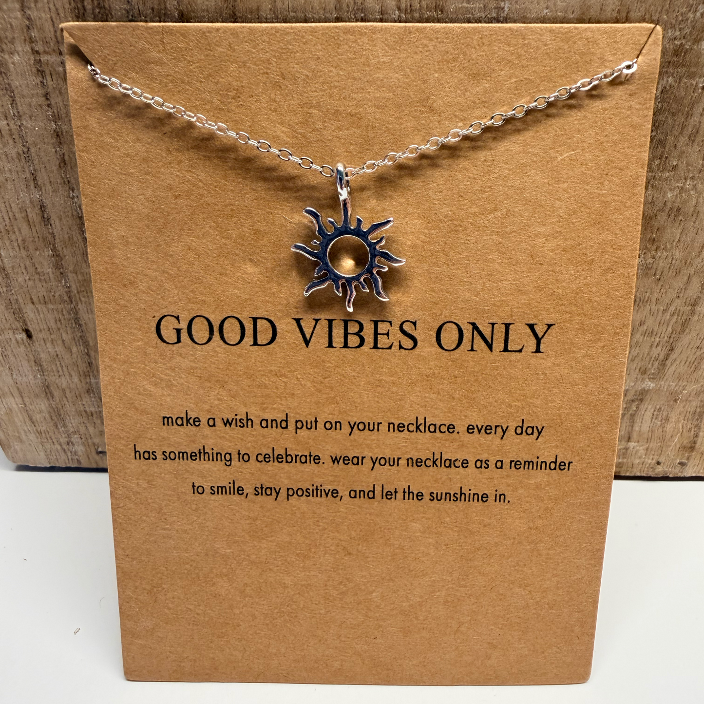 Necklace with sun pendant on a brown card with 'Good Vibes Only' text, placed on a wooden surface.