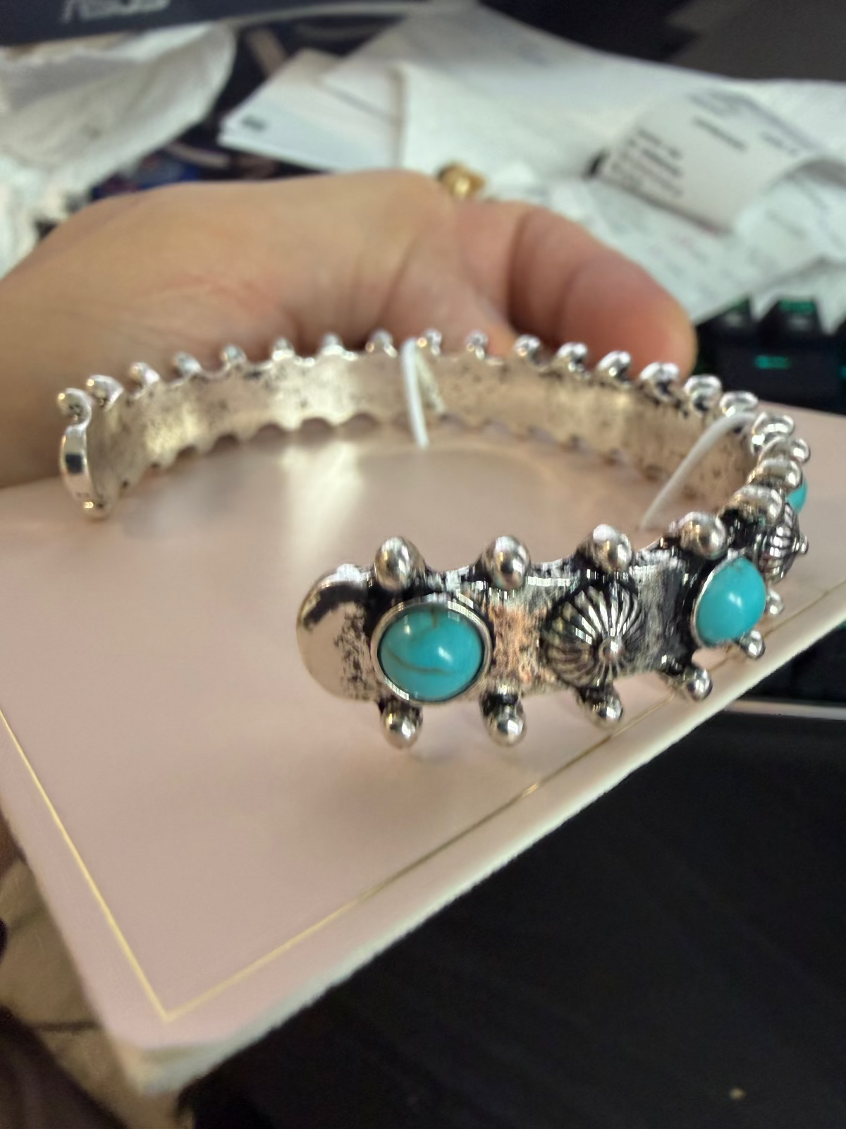 A sterling silver cuff bracelet with turquoise stone accents, held in a hand over a light pink jewelry box.