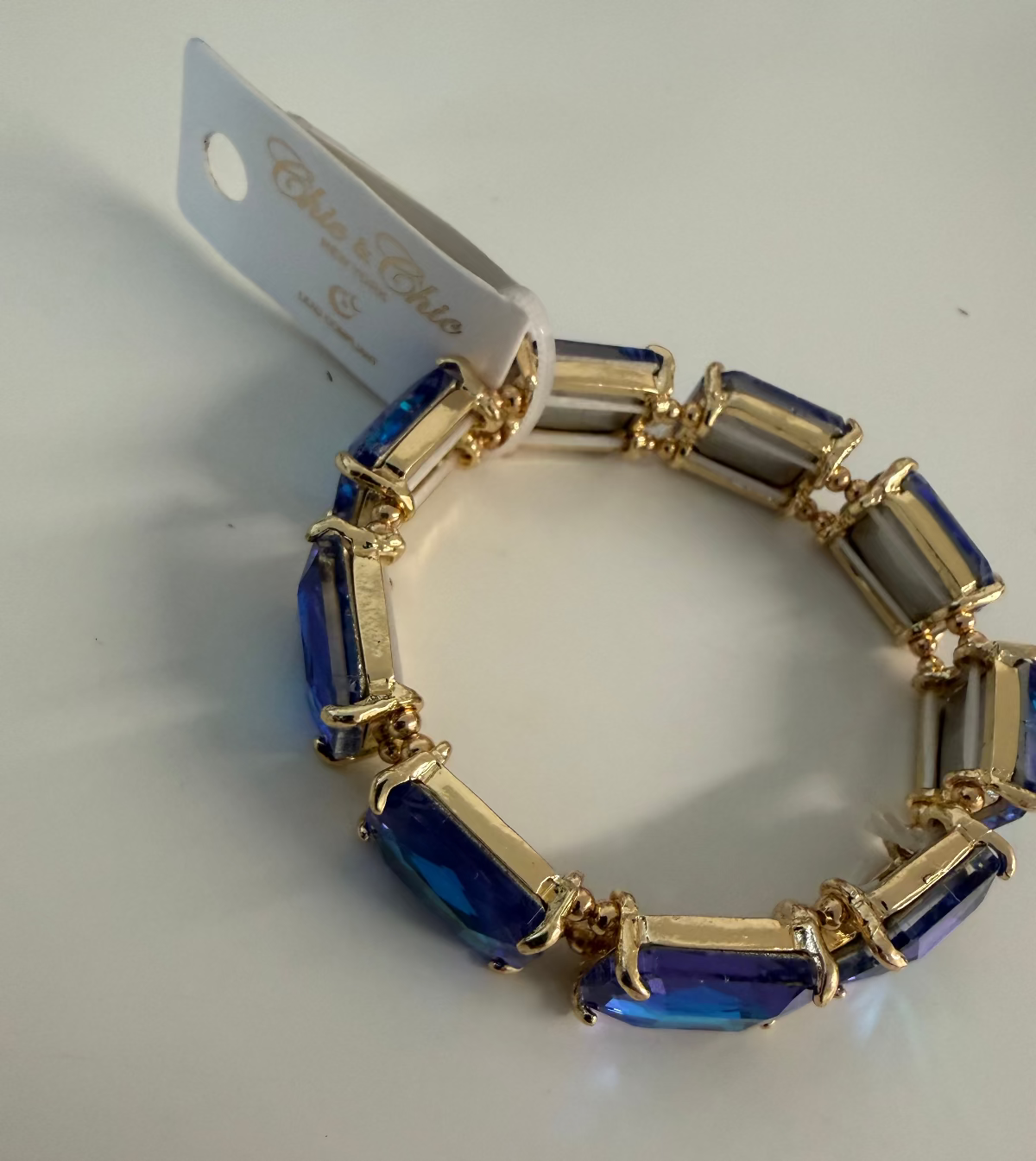 A gold-plated stretch bracelet featuring rectangular blue faceted crystals, shown with its white paper tag attached on a plain white surface.