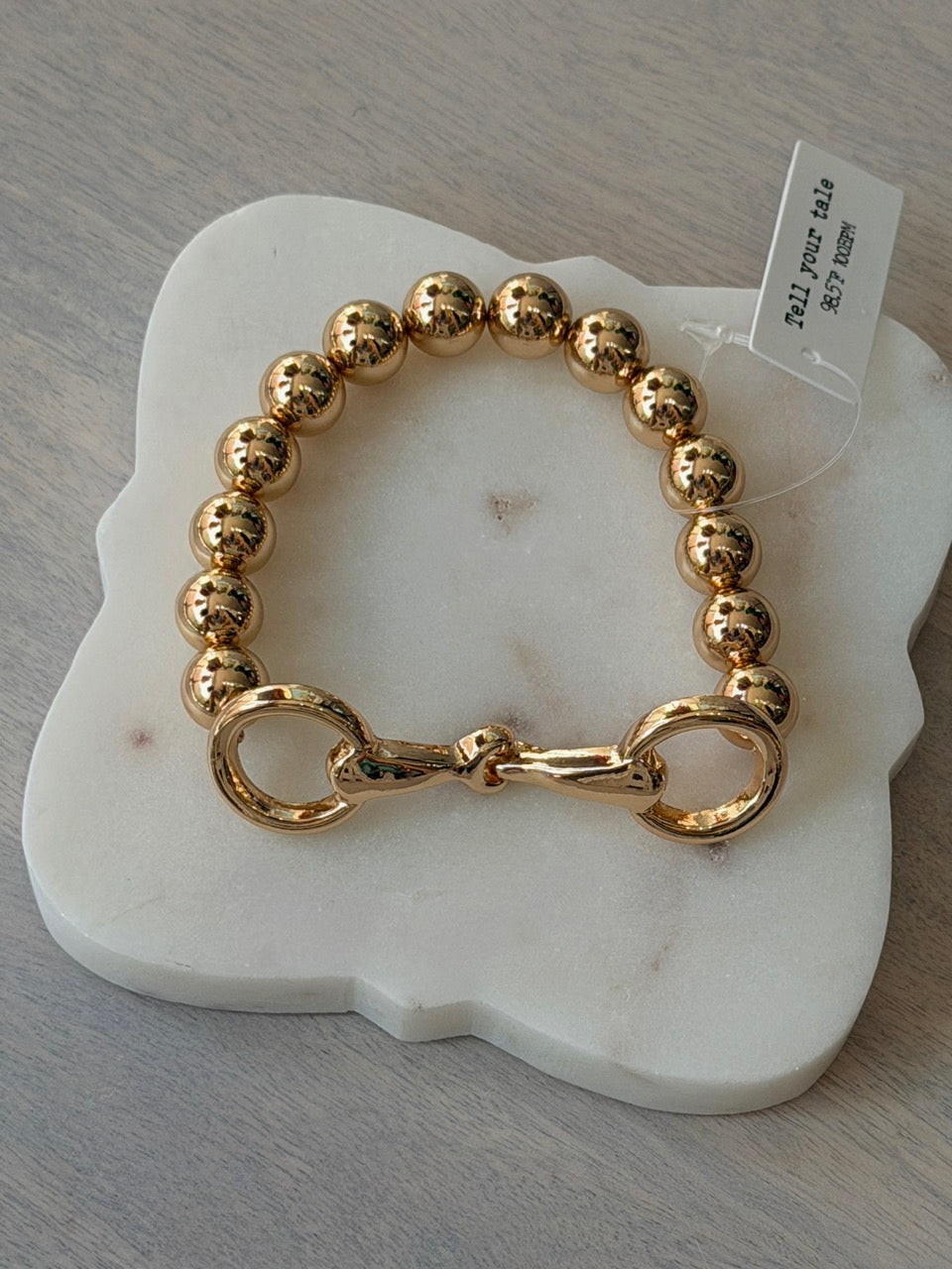 A gold beaded bracelet with a lobster clasp, displayed on a white marble stand over a gray textured surface.