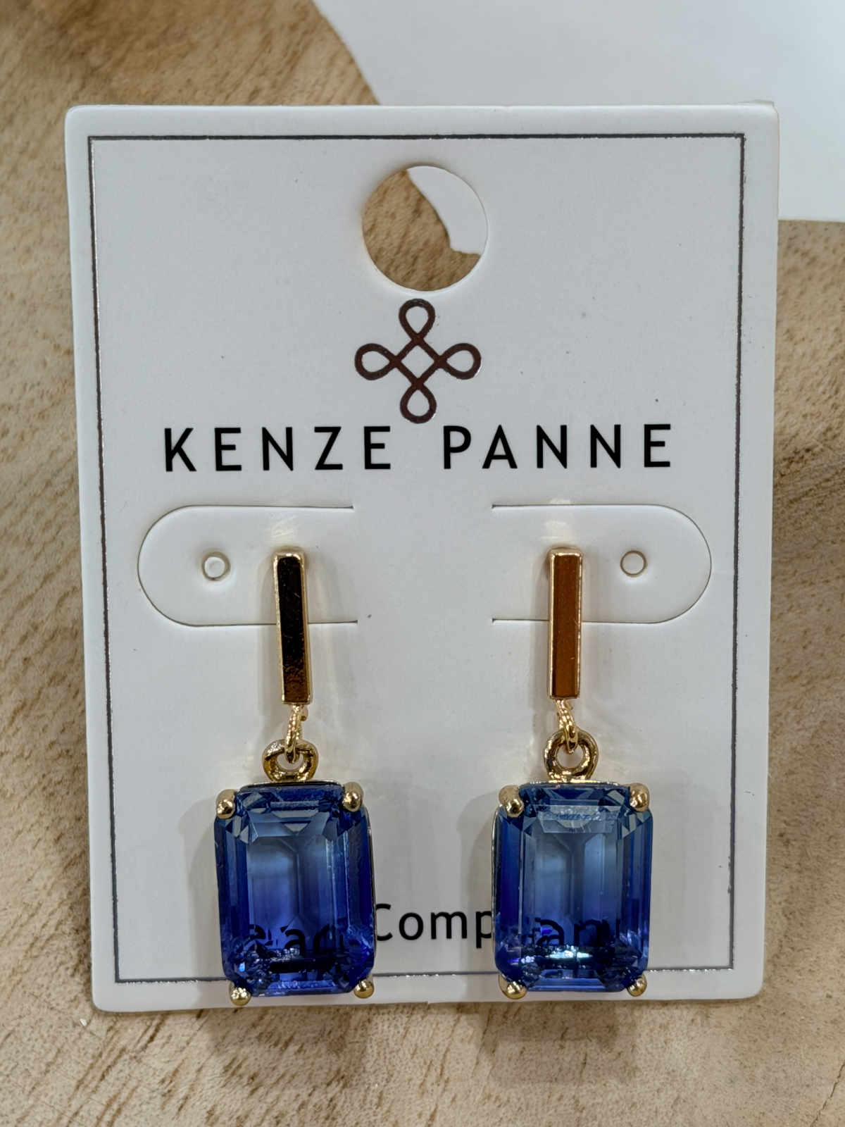 A pair of Kenze Panne drop earrings with rectangular blue gemstones in gold settings, displayed on a white branded card against a light wood background.
