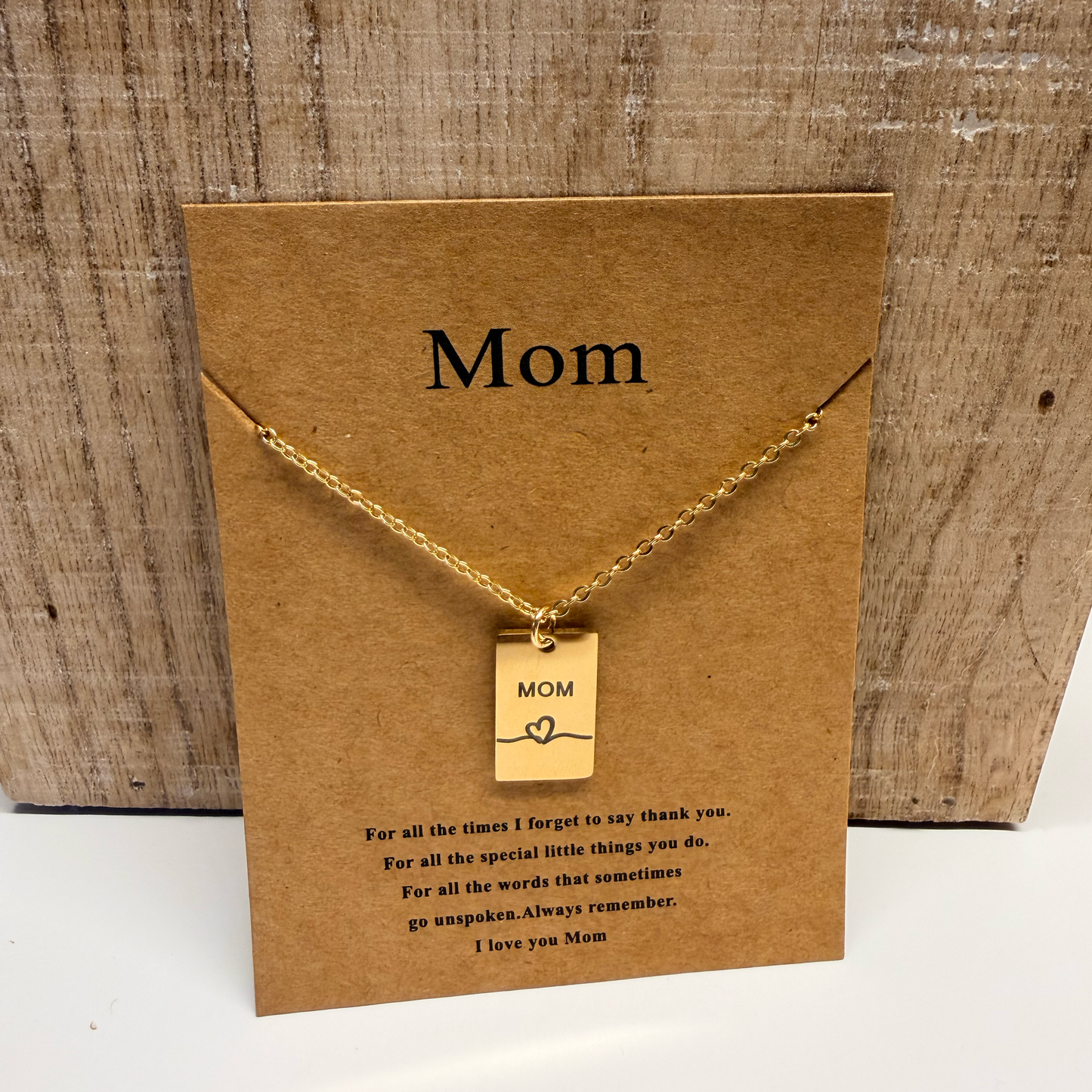 Gold necklace with 'Mom' tag on a brown card with 'Mom' text, on a wooden surface.