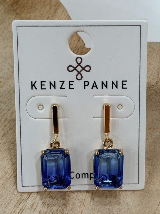 A pair of Kenze Panne drop earrings with rectangular blue gemstones in gold settings, displayed on a white branded card against a light wood background.