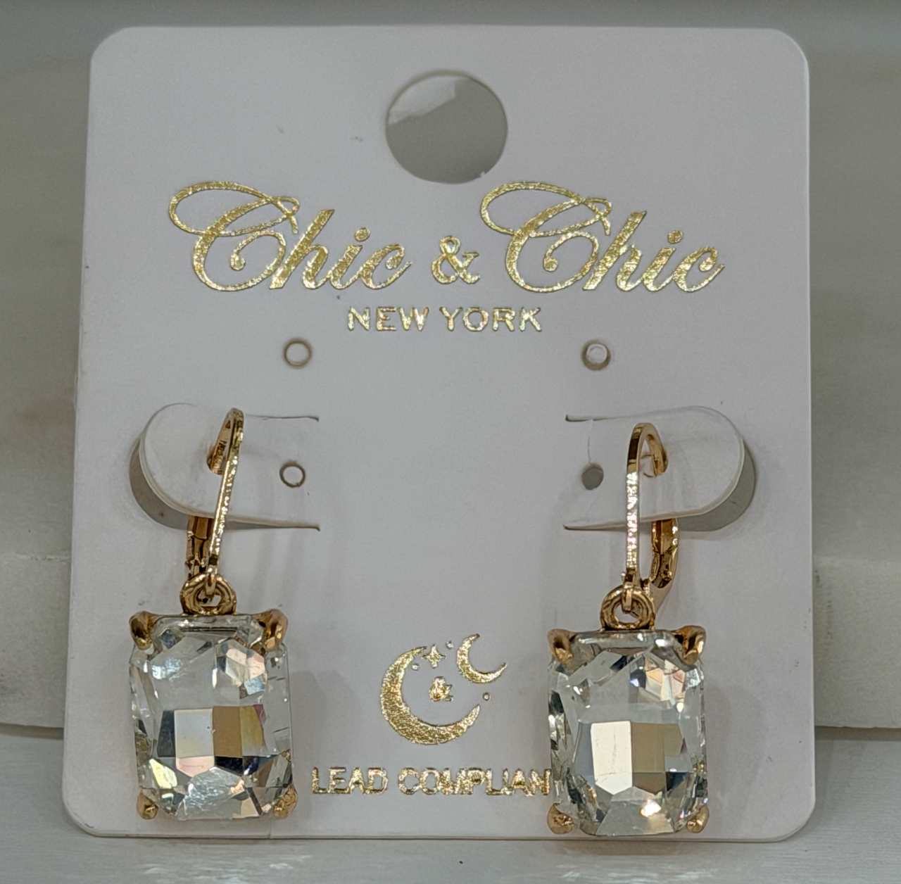 A pair of gold leverback earrings with rectangular clear faceted crystals, displayed on a white 'Chic & Chic New York' branded card.