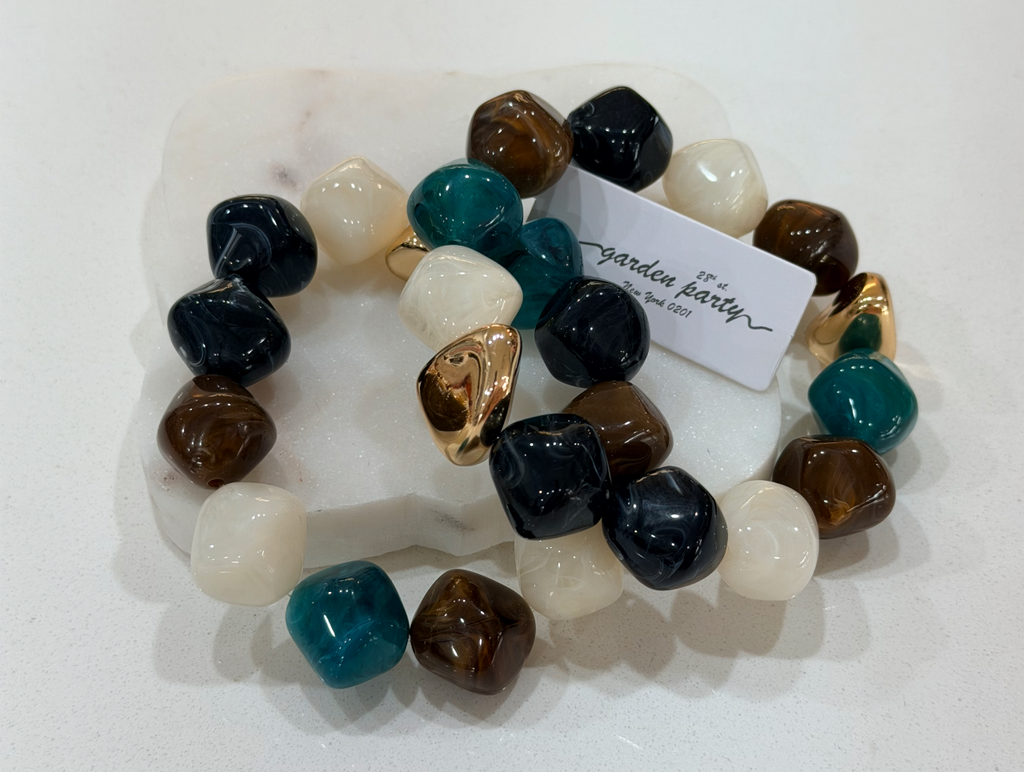 A beaded stretch bracelet displayed on a white marble stand. The bracelet features glossy, polished tumbled stones in white, dark navy, teal blue, and brown, with a small gold heart charm.
