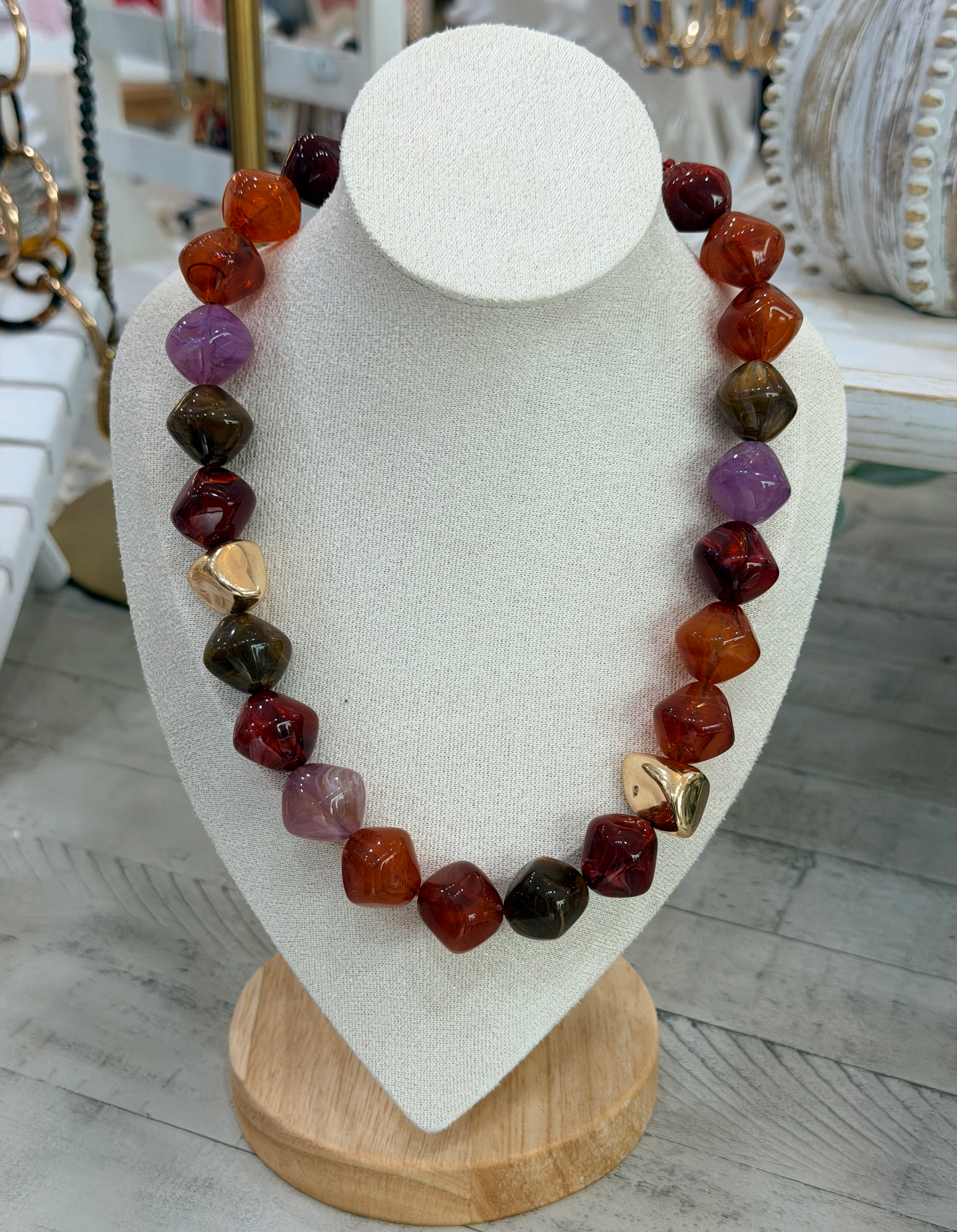 A beaded statement necklace made of faceted gemstone beads in orange, red, purple, and brown, accented with two gold heart charms, displayed on a white jewelry bust.