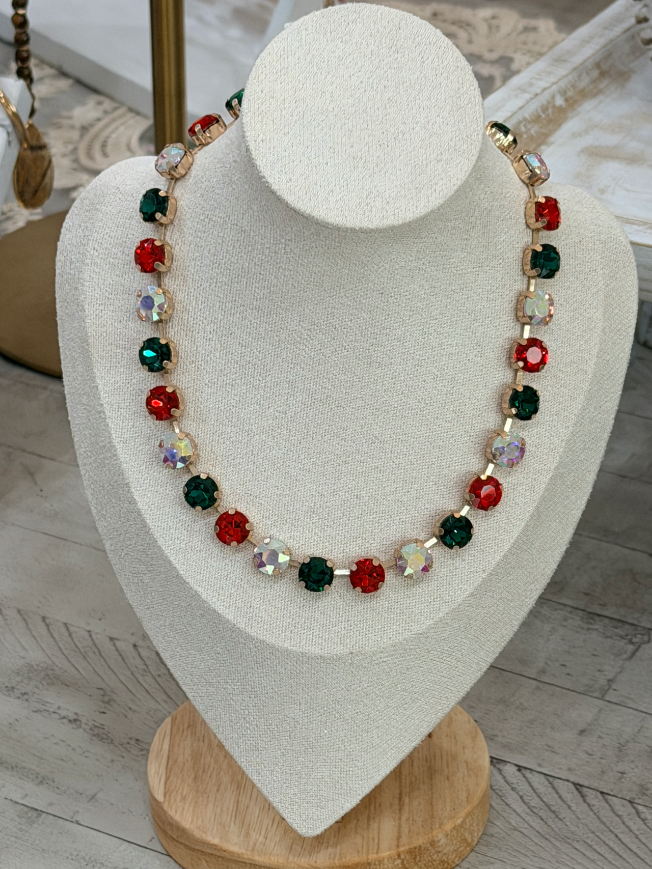 A festive holiday statement necklace with red, green, and iridescent faceted gemstones in gold-toned settings, displayed on a cream-colored jewelry bust.
