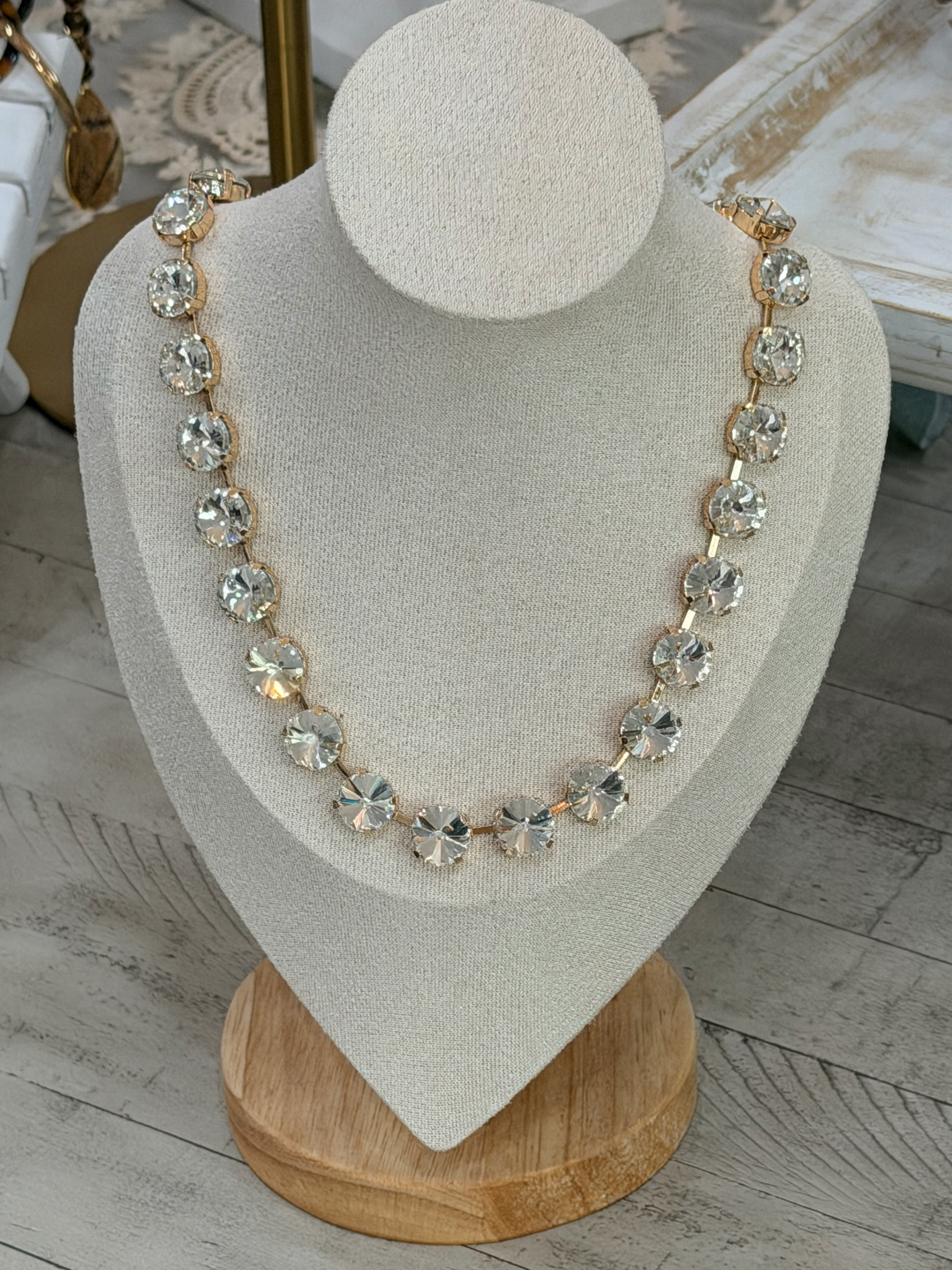 A gold-tone statement necklace with clear faceted rhinestones displayed on a beige fabric jewelry bust stand.