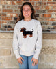 A smiling woman stands against a brick wall wearing a light heather gray sweatshirt with a brown dachshund wearing a cowboy hat graphic on the front, paired with blue jeans.
