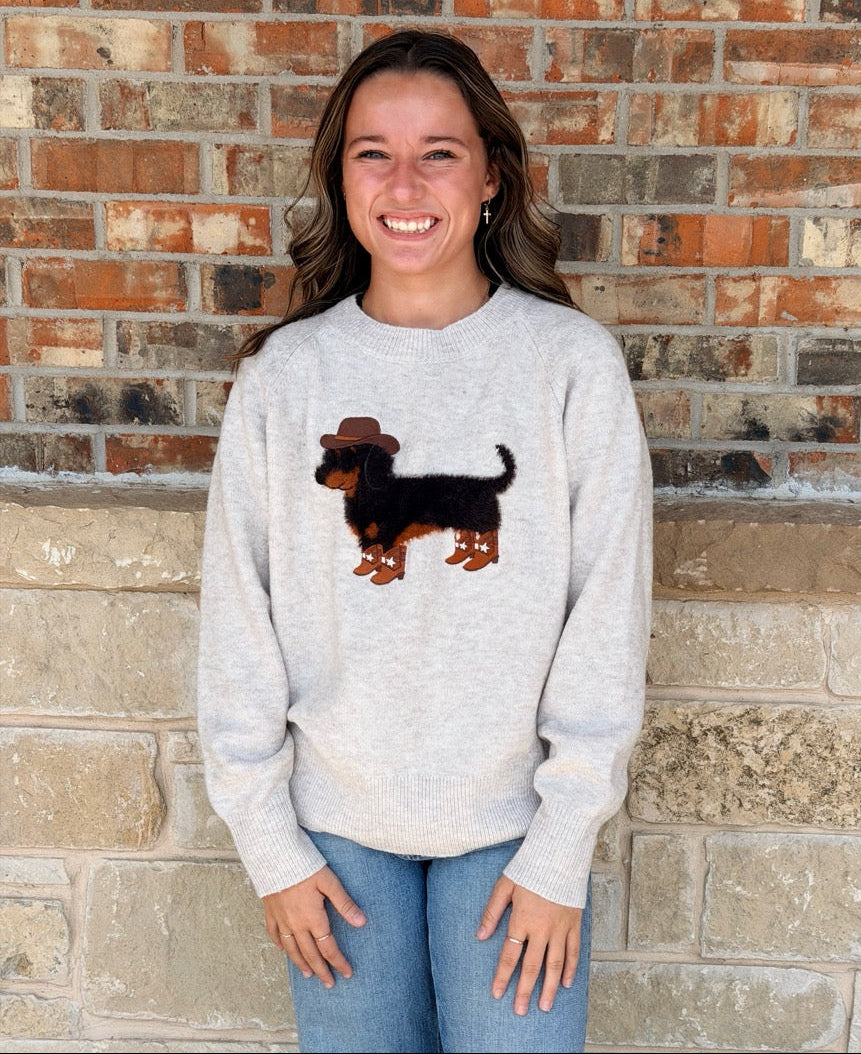 A smiling woman stands against a brick wall wearing a light heather gray sweatshirt with a brown dachshund wearing a cowboy hat graphic on the front, paired with blue jeans.