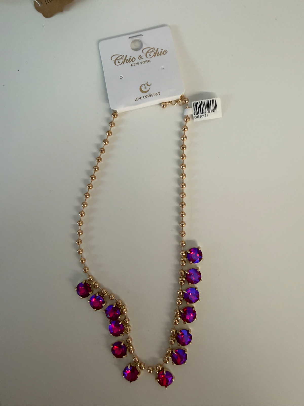 A gold-tone beaded necklace with a V-shaped drop of faceted purple and magenta rhinestones, attached to a white 'Chic & Chloé New York' retail display card, photographed on a light off-white surface.