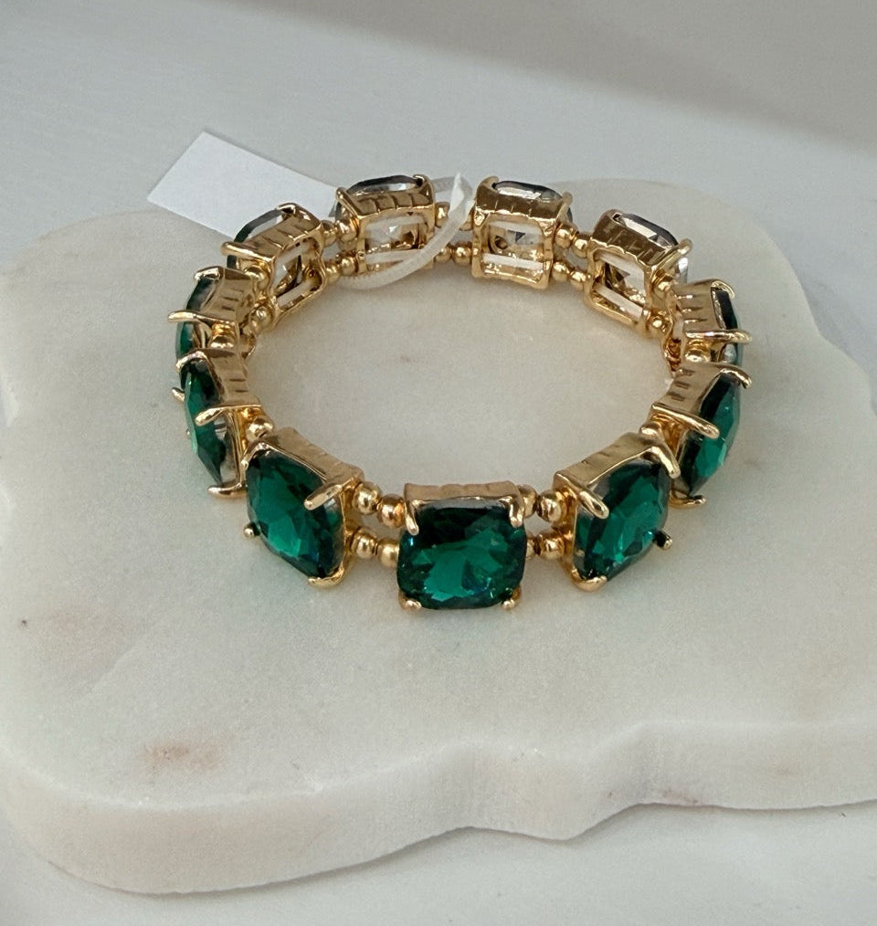A gold-plated bracelet featuring large, square-cut emerald green gemstones, displayed on a white stone stand.
